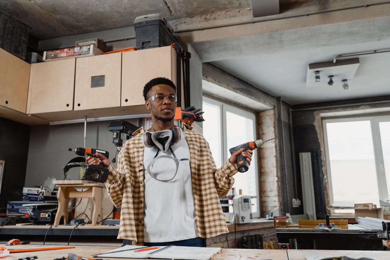 A young craftsman working in a well-equipped woodworking shop, holding power drills.